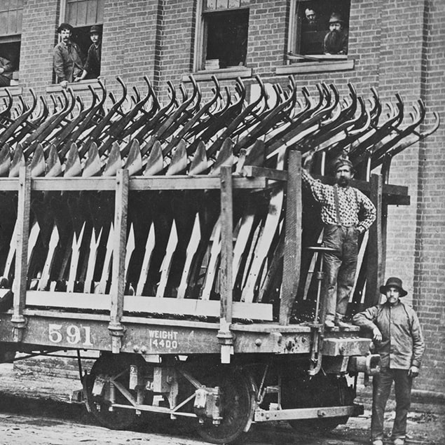 Fotografie z roku 1882, na které jsou tři muži stojící vedle železničního vagónu opatřeného nápisem Deere & Co a naloženého ocelovými pluhy připravenými k odeslání, zatímco z oken budovy v pozadí vyhlížejí zaměstnanci továrny