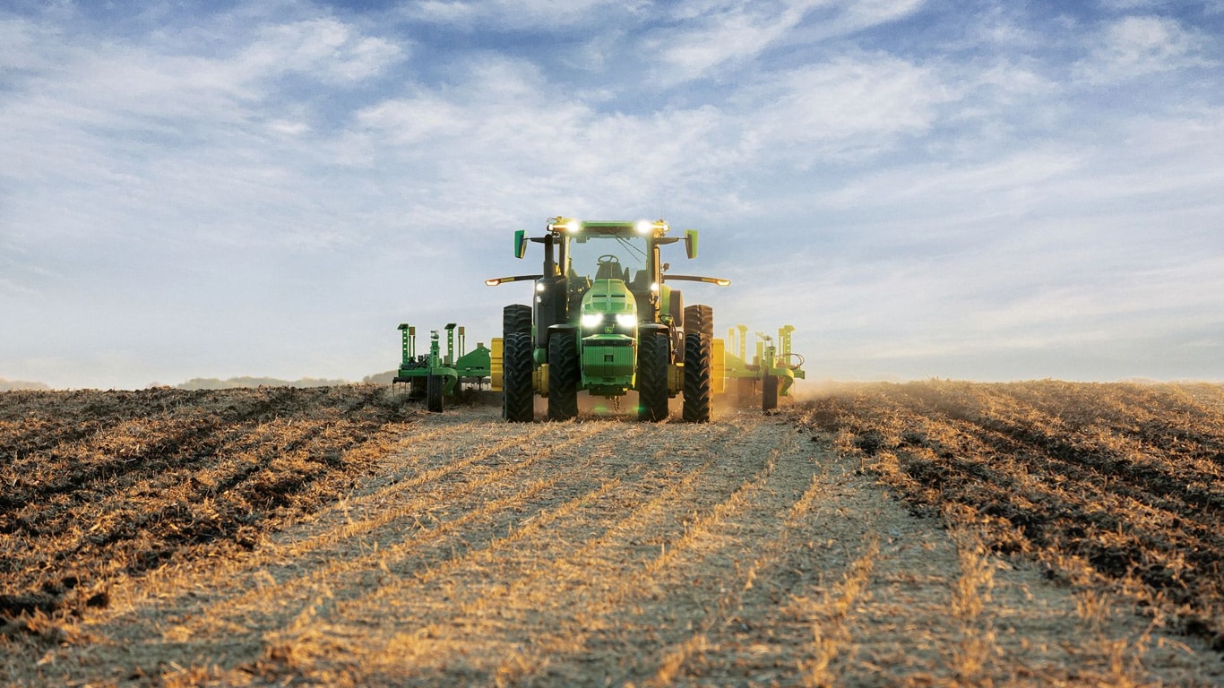 Samojízdné traktory John Deere táhnou zařízení pro obdělávání půdy na otevřené ploše.