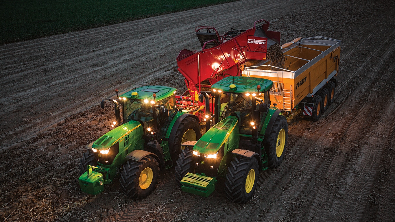 Traktor v&nbsp;noci t&aacute;hne stroj na&nbsp;sklizeň brambor a&nbsp;vykl&aacute;d&aacute; brambory do&nbsp;př&iacute;věsu tažen&eacute;ho jin&yacute;m traktorem