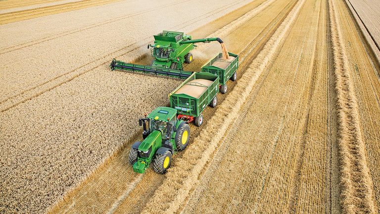 Skl&iacute;zec&iacute; ml&aacute;tička John Deere řady S vykl&aacute;d&aacute; p&scaron;enici do&nbsp;dvou př&iacute;věsů tažen&yacute;ch traktorem