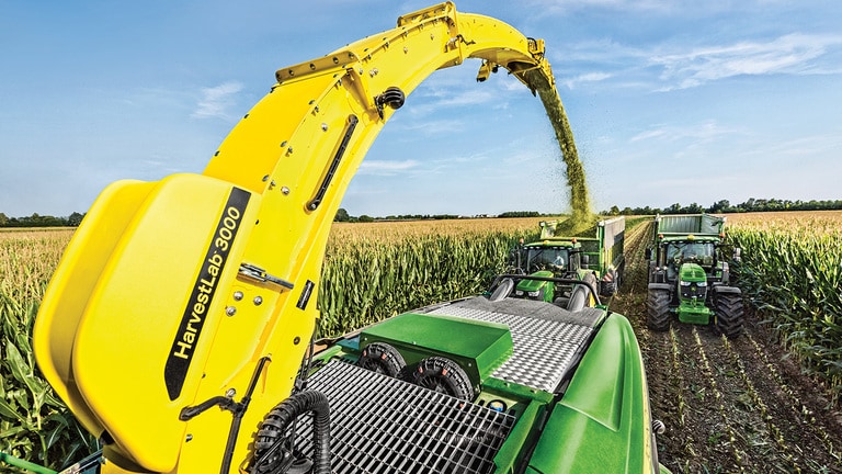 Pohled zezadu na&nbsp;samoj&iacute;zdnou skl&iacute;zec&iacute; řezačku John Deere řady 9000 se&nbsp;dvěma sil&aacute;žn&iacute;mi př&iacute;věsy tažen&yacute;mi traktory John Deere jedouc&iacute;mi za&nbsp;n&iacute;