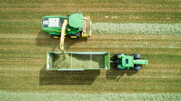 Leteck&yacute; pohled na&nbsp;samoj&iacute;zdnou skl&iacute;zec&iacute; řezačku John Deere řady 8000 se&nbsp;sběračem tr&aacute;vy vykl&aacute;d&aacute; sil&aacute;ž do&nbsp;př&iacute;věsu tažen&eacute;ho traktorem John Deere