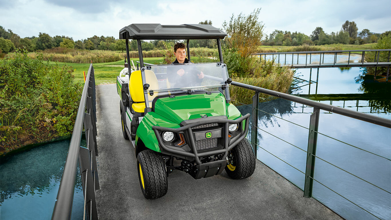 Užitkové vozidlo John Deere Gator GS projíždí po úzkém mostě nad klidným vodním tokem, obklopené bujnou zelení a stromy.