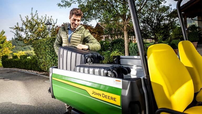 Osoba nakládající velký černý kufr na korbu vozidla John Deere Gator GS s jasně žlutými sedadly, obklopená zelení a stromy.