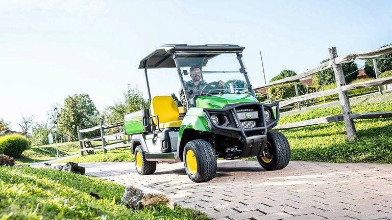 Zelené užitkové vozidlo John Deere Gator GS jedoucí po zpevněné cestě uprostřed upravené krajiny s trávníkem a dřevěnými ploty.