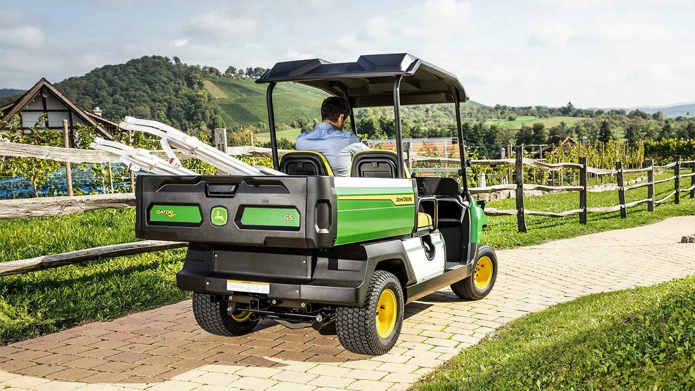 Zadní pohled na vozidlo John Deere Gator GS jedoucí po cihlové cestě s malebnými kopci a vinicemi v pozadí.