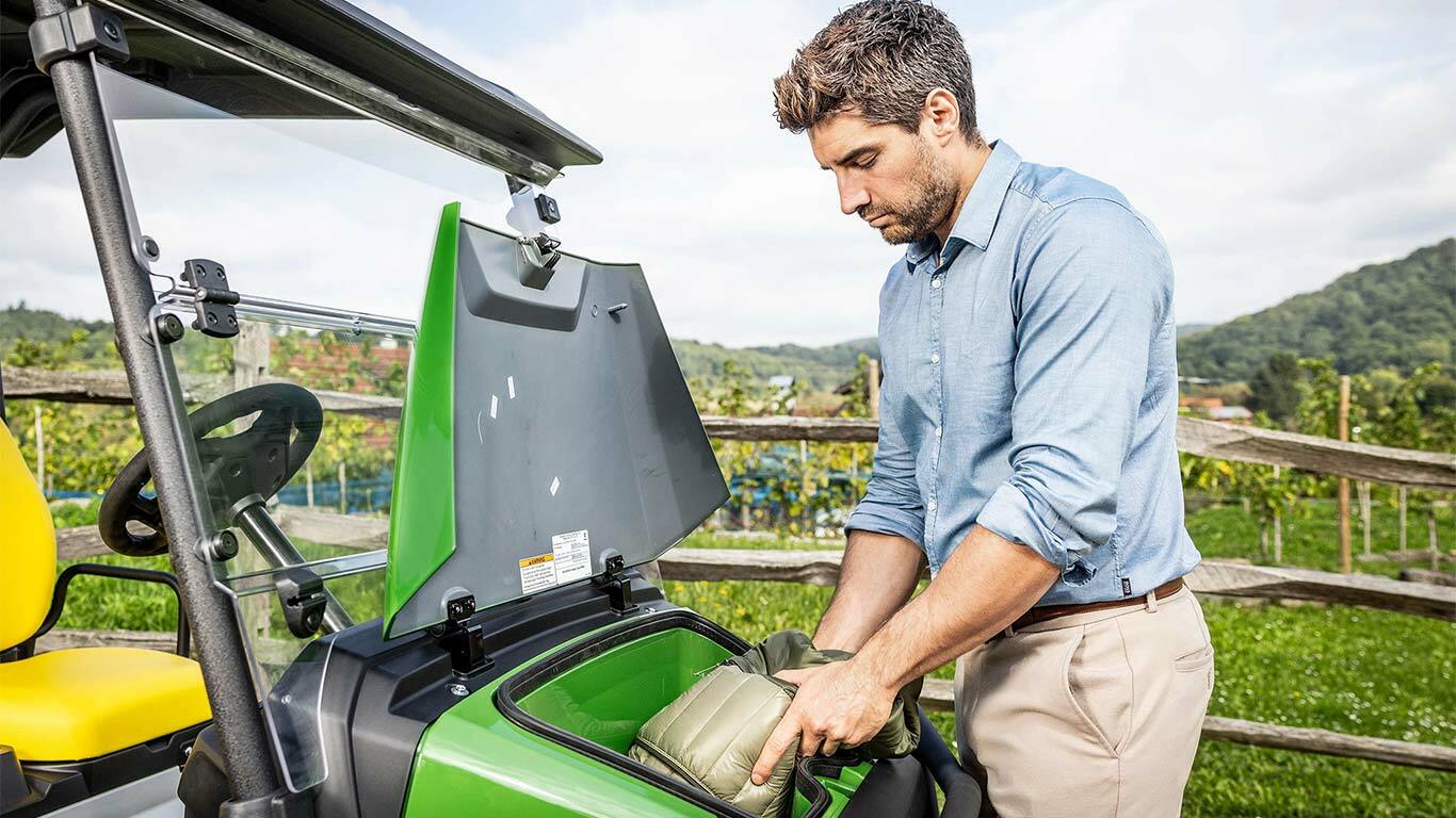 Osoba ukládající polstrovanou tašku do předního úložného prostoru zeleného vozidla John Deere Gator GS zaparkovaného venku poblíž dřevěného plotu a vinice.
