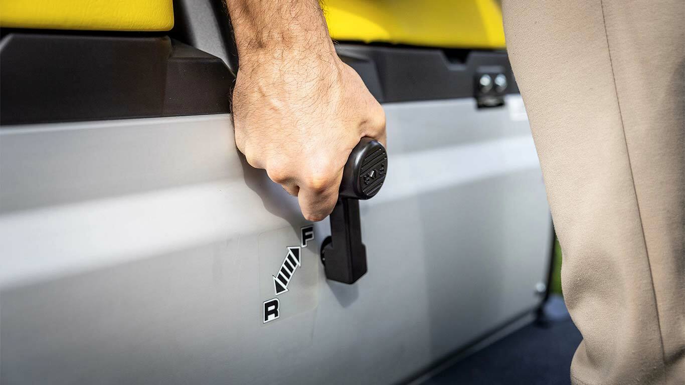 Detailní záběr ruky ovládající řadicí páku na vozidle John Deere Gator GS, znázorňující polohy pro jízdu vpřed a vzad vedle žlutých sedadel.
