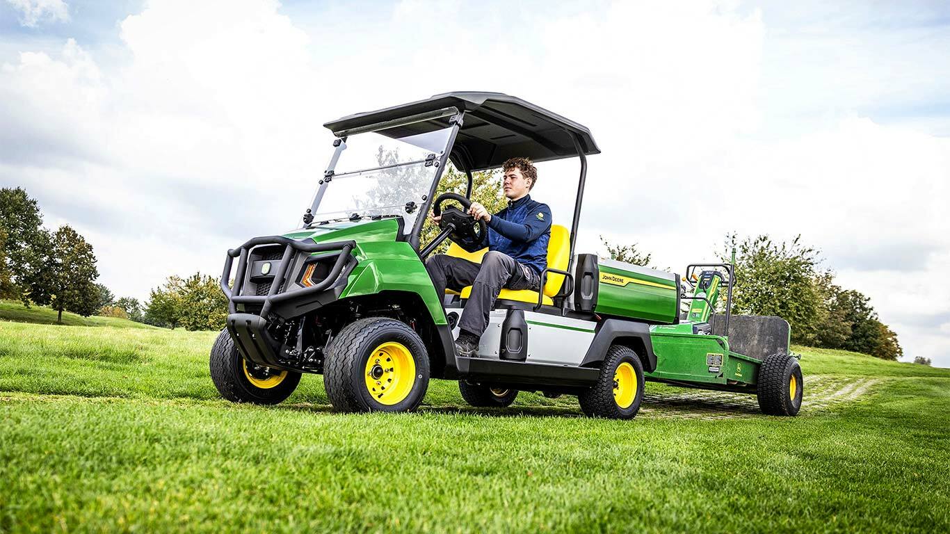 Zelené elektrické užitkové vozidlo John Deere Gator táhne zařízení přes travnaté pole pod částečně zataženou oblohou.