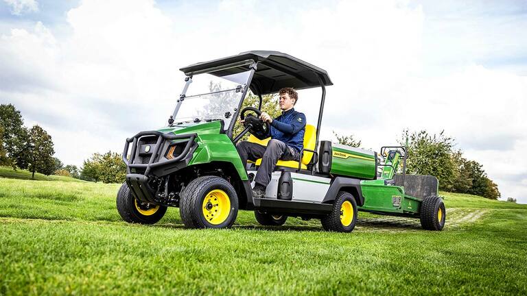 Zelené elektrické užitkové vozidlo John Deere Gator táhne zařízení přes travnaté pole pod částečně zataženou oblohou.