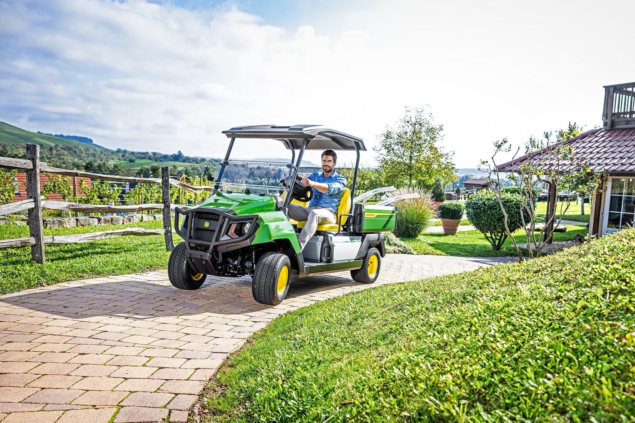 John Deere Gator GS při jízdě po zpevněné cestě poblíž domu