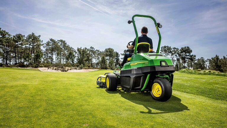 Člověk řídí elektrickou sekačku greenů John Deere E-Cut™ 2775 na dobře udržovaném golfovém hřišti, v pozadí jsou stromy a částečně zatažená obloha.