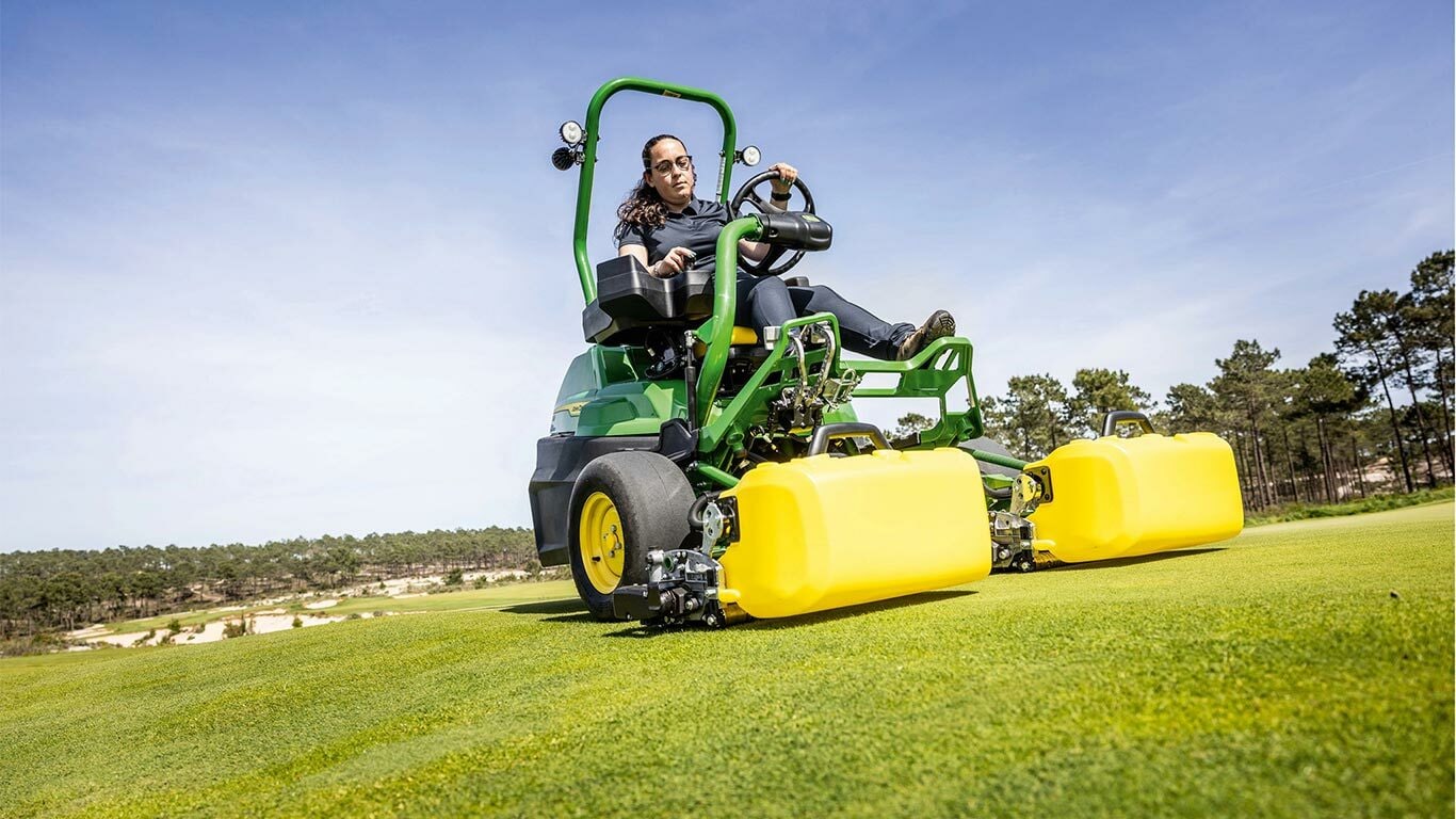 Člověk pracuje se zeleno-žlutou elektrickou sekačkou John Deere E-Cut™ 2775 s předními válci zakrytými žlutým krytem, jede na golfovém greenu, v pozadí jsou stromy a jasně modrá obloha.