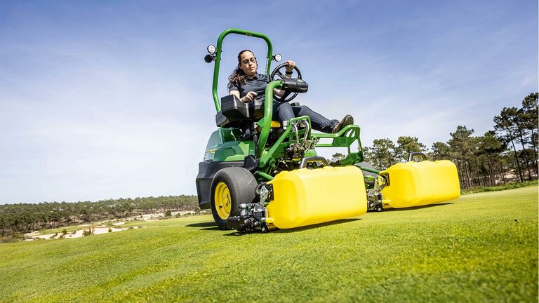Člověk pracuje se zeleno-žlutou elektrickou sekačkou John Deere E-Cut™ 2775 s předními válci zakrytými žlutým krytem, jede na golfovém greenu, v pozadí jsou stromy a jasně modrá obloha.