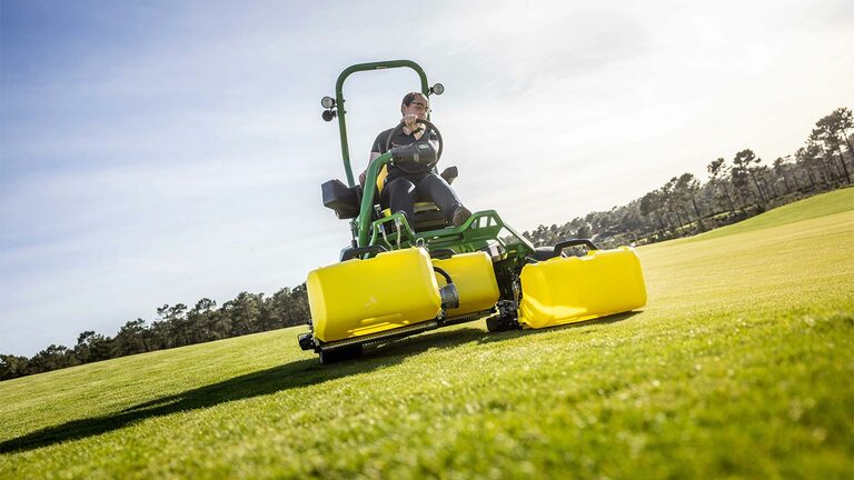 Člověk pracuje se zeleno-žlutou elektrickou sekačkou John Deere E-Cut™ 2775 se třemi velkými předními válci zakrytými žlutými kryty, jede na golfovém greenu, v pozadí jsou stromy a částečně zatažená obloha.