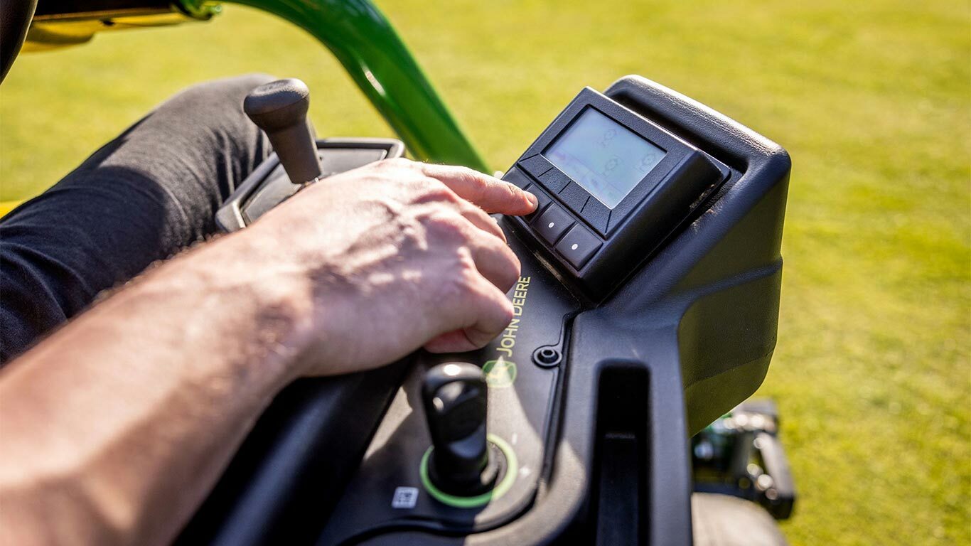 Detailní záběr ruky osoby, která používá ovládací panel elektrické sekačky John Deere E-Cut™ 2775, mačká tlačítka na digitálním displeji, kde je vidět joystick a další ovládací prvky.