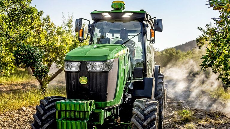 Zelenožlutý traktor John Deere 5075EN projíždí ovocným sadem po polní cestě, obklopený ovocnými stromy a slunečním světlem.