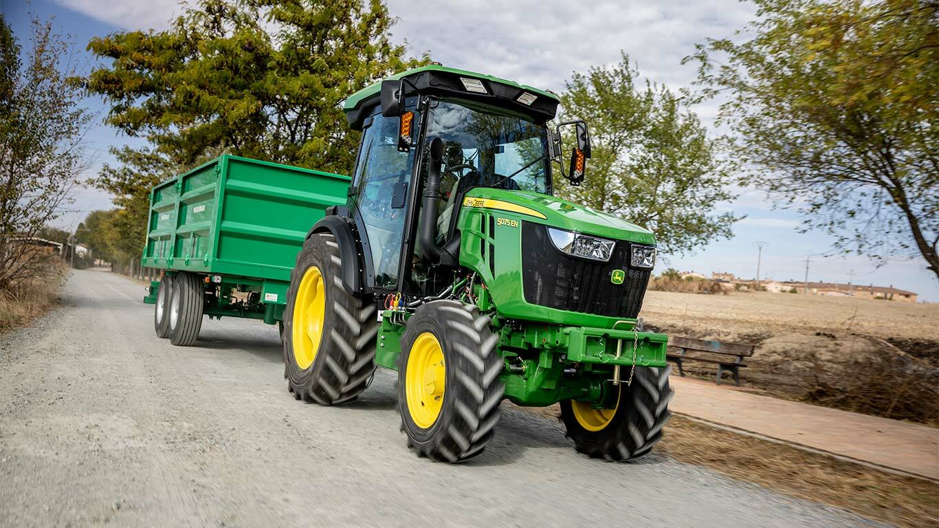 Traktor John Deere 5075EN táhne zelený přívěs po venkovské polní cestě s stromy v pozadí.