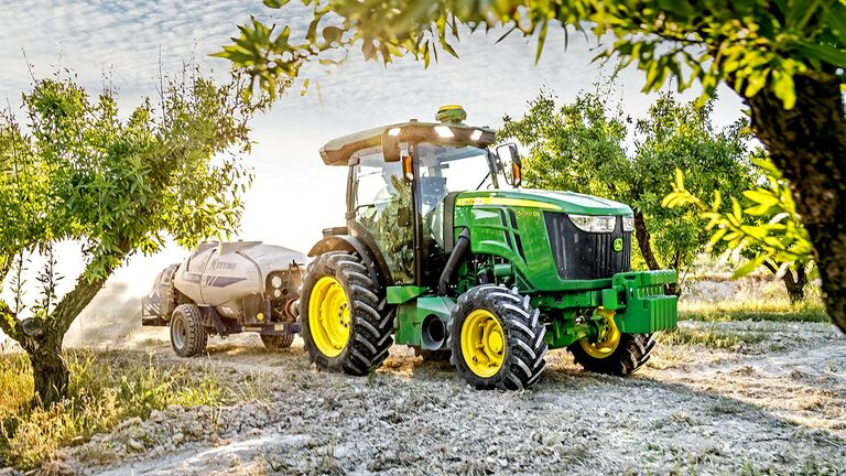 Zelenožlutý traktor John Deere 5090EN táhne postřikovač sadem za slunečného dne.