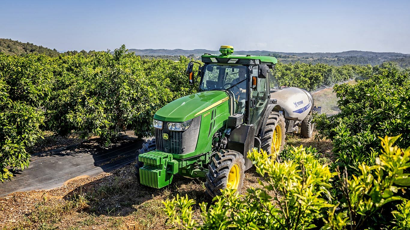 Zelenožlutý traktor John Deere 5105EN táhne postřikovač v rozkvetlém sadu s kopcovitou krajinou v pozadí.