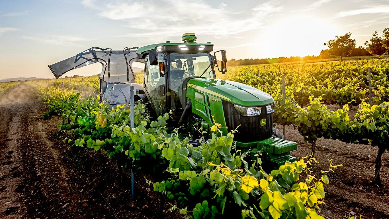 Zelenožlutý traktor John Deere 5105EN pracuje ve vinici při západu slunce a postřikuje rostliny mezi řádky vinné révy.