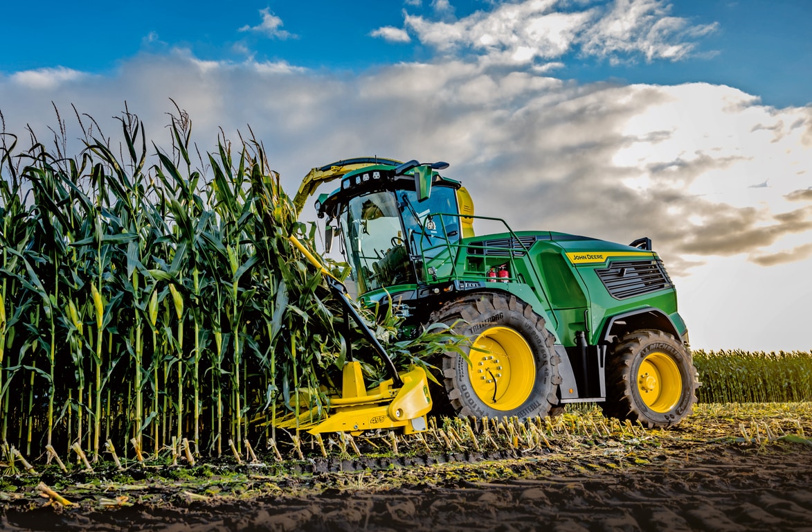 Sklízecí řezačka John Deere sklízející kukuřici na poli pod modrou oblohou s mraky.