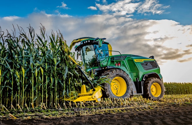 Sklízecí řezačka John Deere sklízející kukuřici na poli pod modrou oblohou s mraky.
