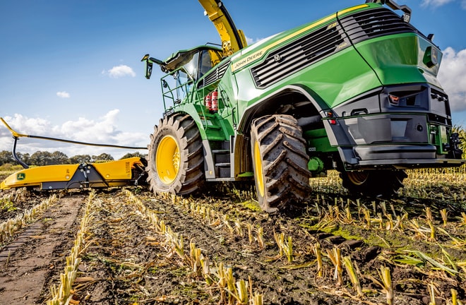Sklízecí řezačka John Deere pracující na sklízeném kukuřičném poli, jsou vidět velké pneumatiky a žací mechanismus stroje na pozadí jasné modré oblohy.