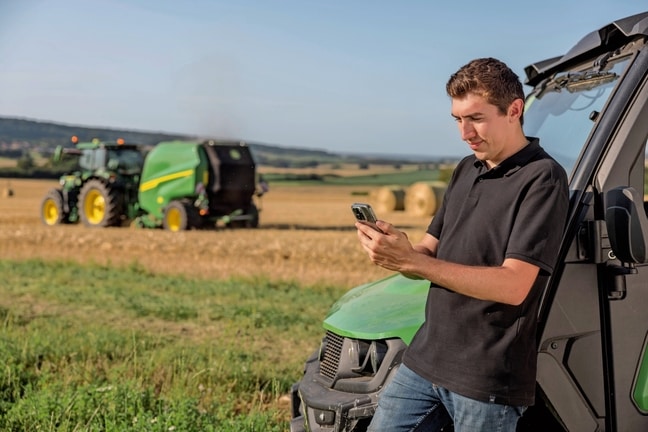 Osoba, která kontroluje data dokumentace balíku v mobilním telefonu se sběracím lisem John Deere v pozadí