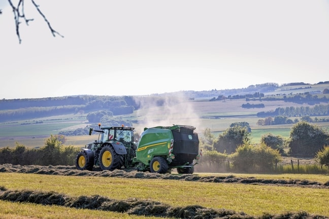 John Deere V462R tažený traktorem John Deere 6R 185 při lisování sena